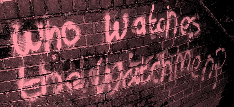 Photo d'un mur sur lequel est écrit "Who whatches the Watchmen?" tagué à la bombe de peinture.